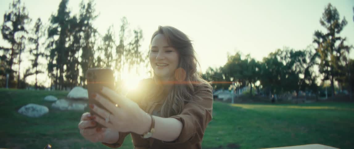 A young woman sits on a bench in a public park at sunset her face glowing with excitement as she chats on a video call the warm hues of the setting sun cast a golden light around her adding to the joyful energy of the moment