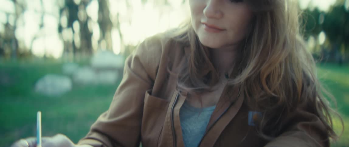 A persons hands are shown up close holding a thin paintbrush over a watercolor palette the scene is outdoors on a sunny day with the person wearing a brown jacket and a watch