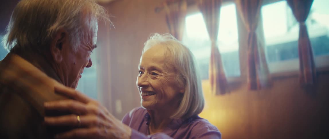 A closeup shot focuses on an elderly woman with white hair wearing a lilac shirt who is laughing while talking to a man offcamera the scene is indoors brightly lit by light filtering through windows with curtains in the background