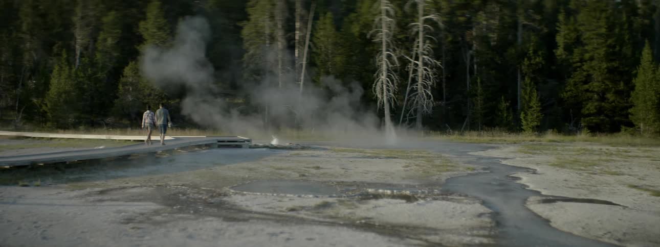 Two people walking in the forest next to steaming water with a couple of small geysers bubbling