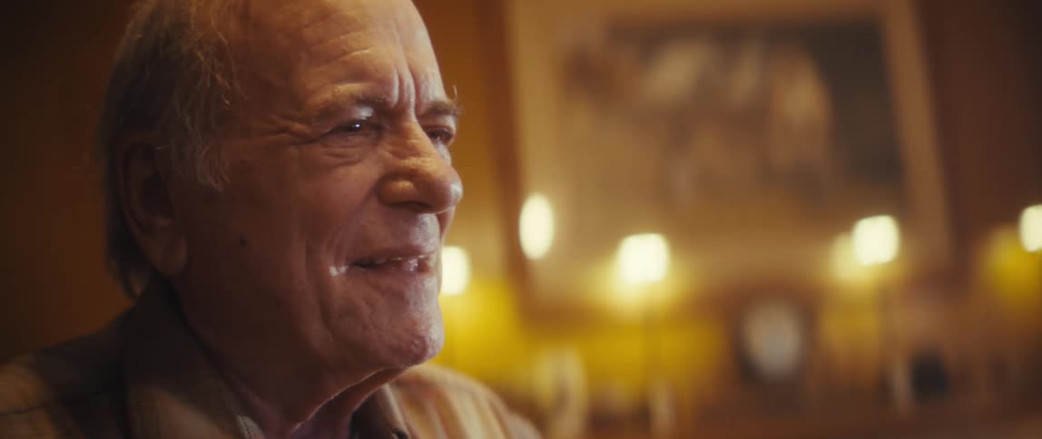 A closeup shot shows an elderly couple brightly lit by warm soft indoor lighting slowdancing and gazing lovingly at each other the man smiles warmly as the woman wearing a lavender shirt tenderly rests her hand on his shoulder