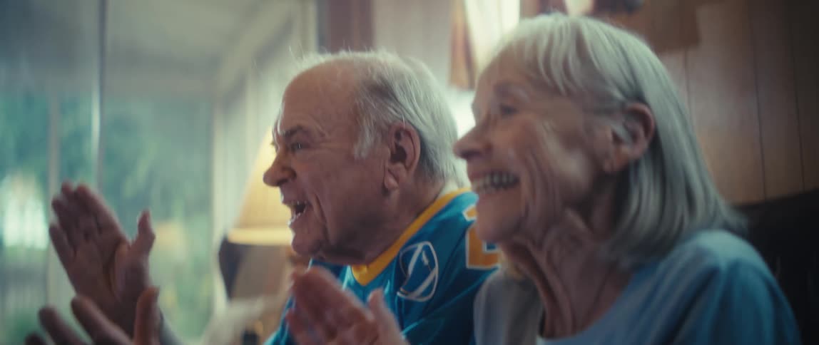 An elderly man wearing a blue and yellow jersey and an elderly woman sit sidebyside yelling and enthusiastically clapping their hands with excited expressions