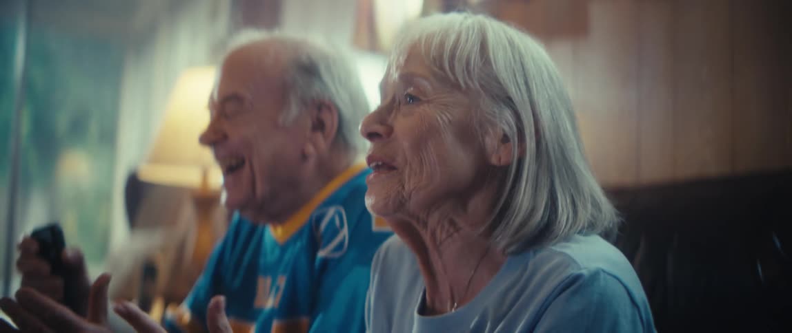 An elderly couple sits sidebyside they clap their hands together with smiles on their faces and they are reacting to an unseen event in an indoor setting