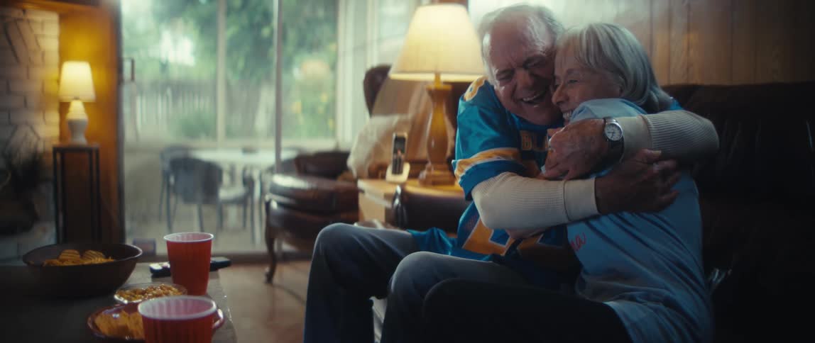 An elderly couple is smiling and hugging on their couch