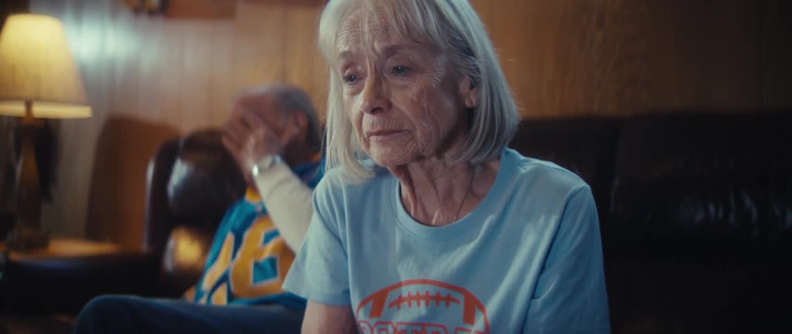 An older woman with white hair is shown in a closeup shot looking slightly down with a serious expression she is seated indoors wearing a light blue tshirt with an orange football graphic and an older man is blurred in the background