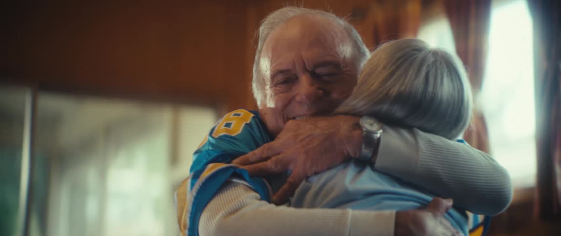 Close up of an elderly couple hugging each other while watching a sports game in a living room during daytime