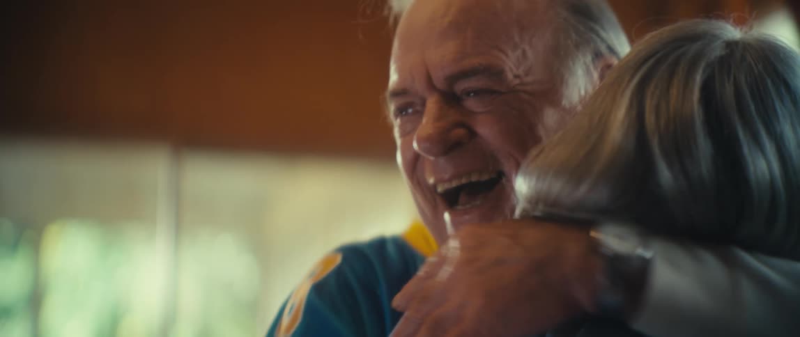 An elderly couple hugs each other excitedly in the living room laughing with joy as they share a heartwarming moment