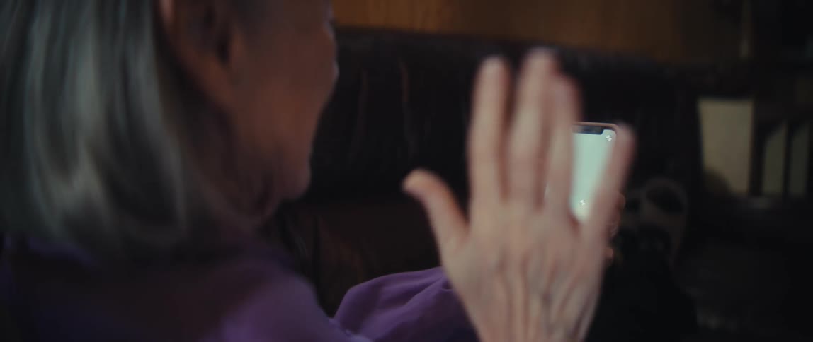 An elderly woman waves at her smartphone which displays a green screen with tracking markers for a video call