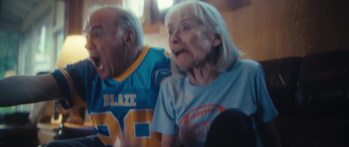 An elderly couple sits leaning forward with worried expressions then both yell in shock before smiling and hugging tightly in celebration while watching something on a screen in a living room