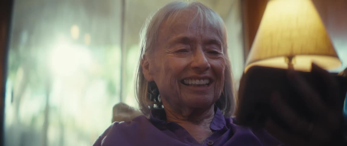 The video is a warm closeup shot of an older woman with white hair sitting indoors initially looking thoughtfulshe then looks toward something offcamera presumably a phone or a person and bursts into a big authentic laugh with a joyful expressio