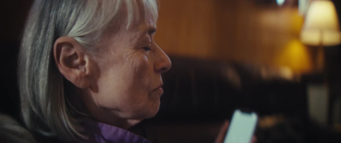 Elderly woman with gray hair holds smartphone with light screen while seated on dark leather couch