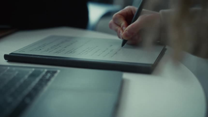 Person writes with digital pen on tablet in workspace with laptop and white table