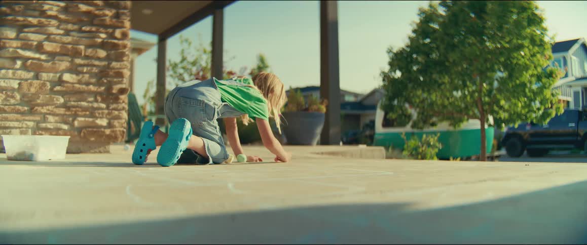 A young girl wearing overalls and bright blue clogs kneels on a sunny patio drawing on the concrete with a piece of white chalk she is focused on her activity with a stone pillar and a residential neighborhood visible in the background