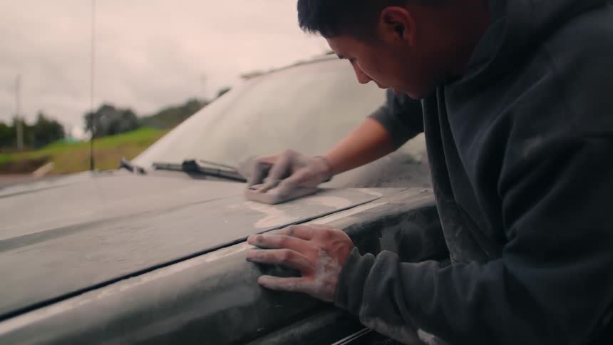 A man wearing a hooded sweatshirt leans over the hood of a vehicle and manually rubs a sanding block against the body to prepare the surface outdoors
