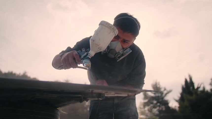 A man wearing a respirator mask and a dark hoodie uses a paint spray gun to refinish a dark surface outdoors under a bright sky