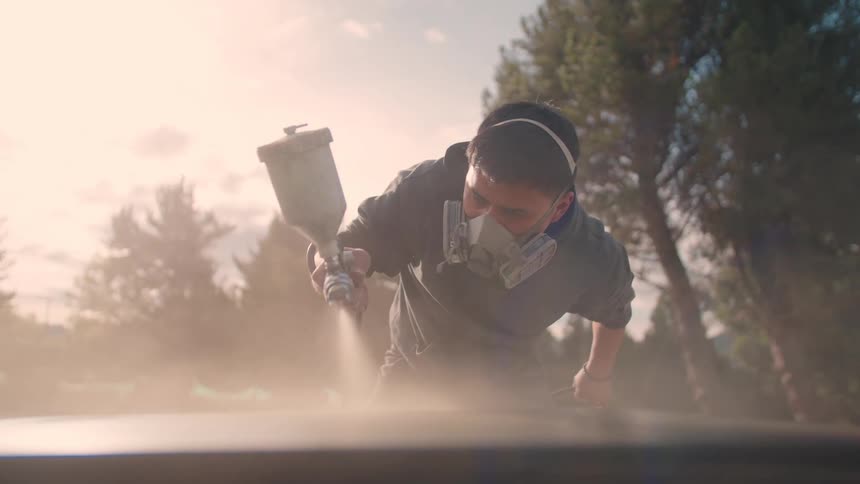 Person performs spray painting on metal surface outdoors using mask and ear protection