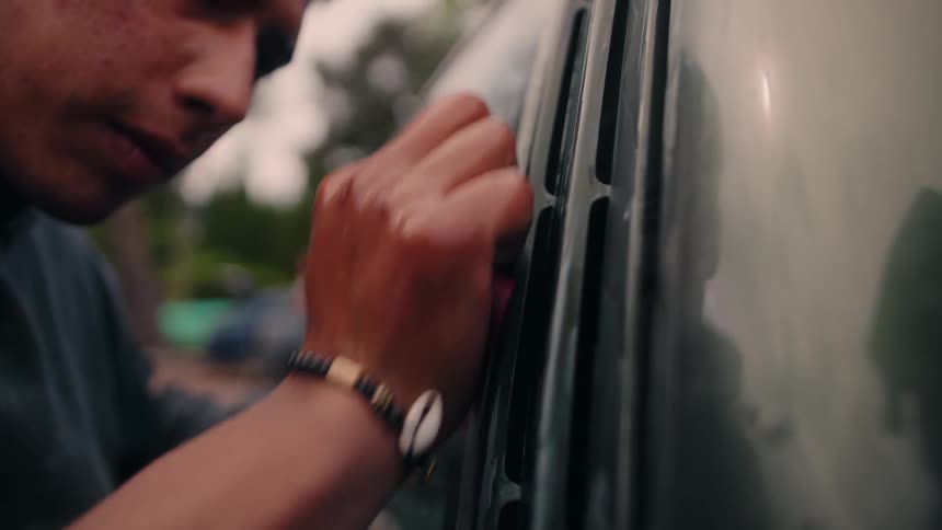 Man uses tool to manipulate car window while wearing shell bracelet in outdoor setting