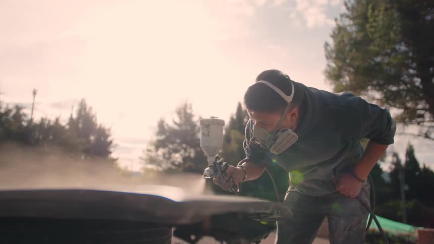 A painter uses spray painting equipment to coat a metal piece at midday focusing on creating a smooth and even finish