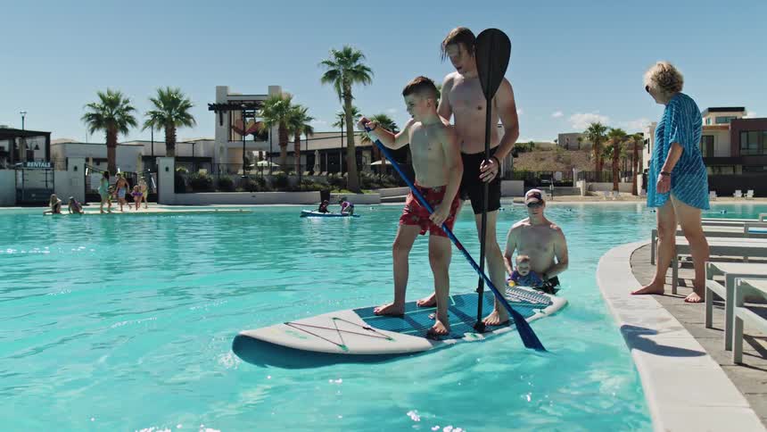 Man and boy paddle together on a board while family members watch by the pool under bright sunlight