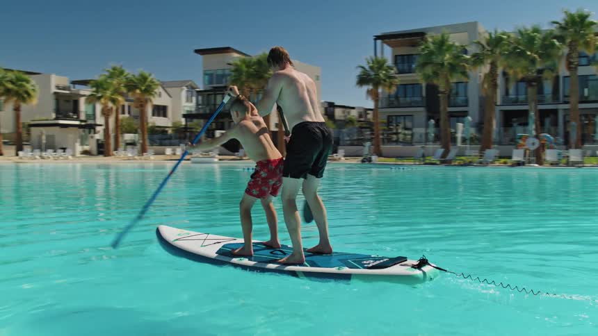 Father and son balance on a paddle board in a large swimming pool and paddle in unison on a summer day