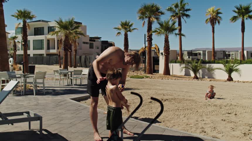 Man applies sunscreen to childs in outdoor area with tables palm trees and buildings in the background