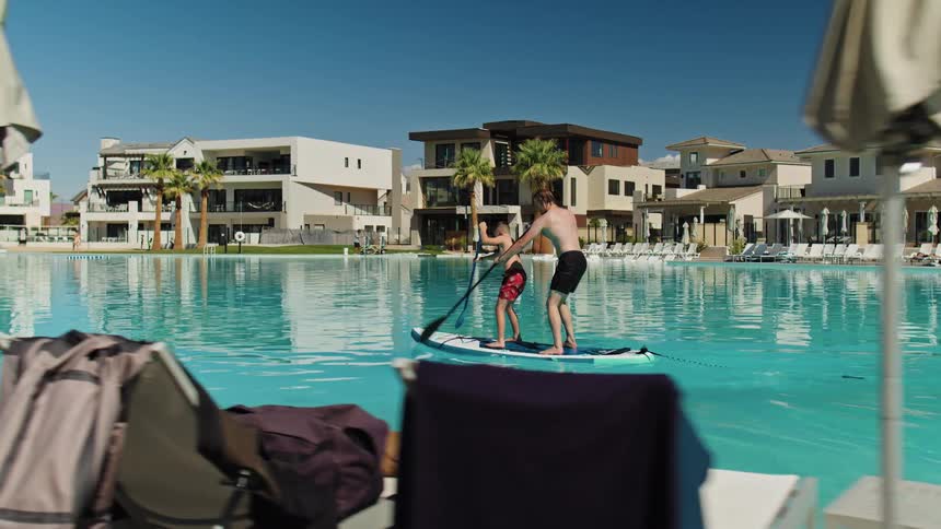 Father and son paddling on paddle board at a pool resort surrounded by villas
