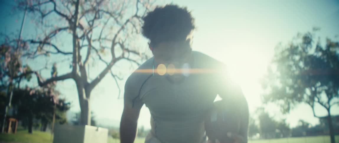 A young athlete backlit by intense sunlight causing a lens flare jogs in place while holding a football in a park