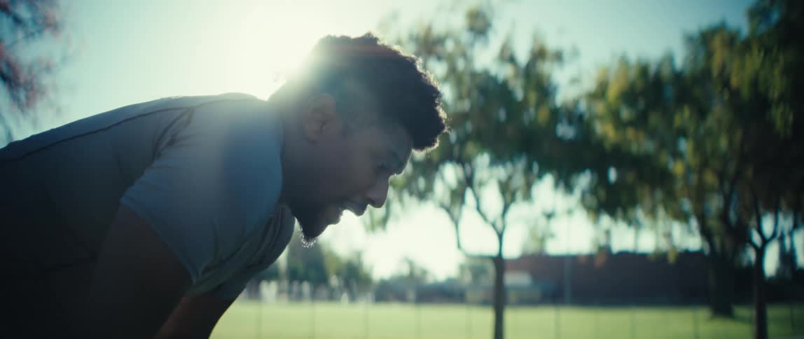 A man with dark curly hair leans forward and breathes heavily after exertion silhouetted against a bright sun in a park setting with trees and a field