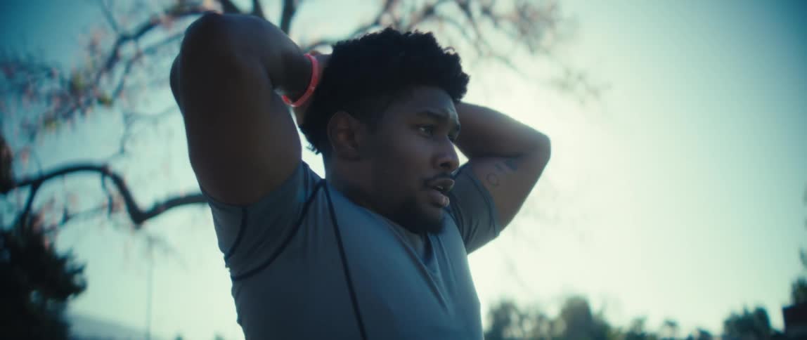 A man wearing a workout shirt stands outdoors looking up before raising his hands to wipe sweat or adjust his hair after a period of exertion the scene is brightly backlit by the sun which flares dramatically around him and a large bare tree