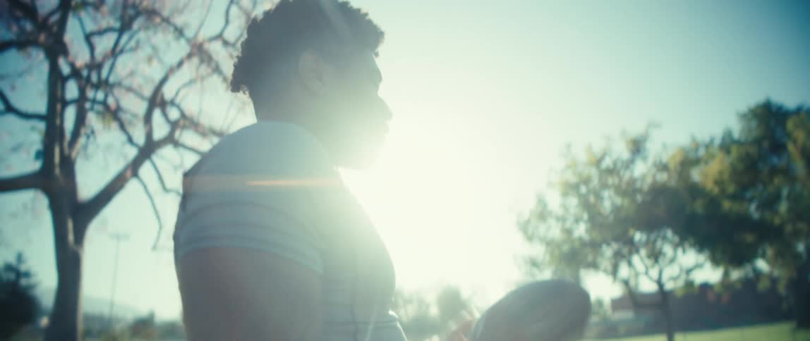 A man in a gray tshirt stands outdoors he holds an american football in his hands and he is silhouetted against the bright sun with trees visible in the background