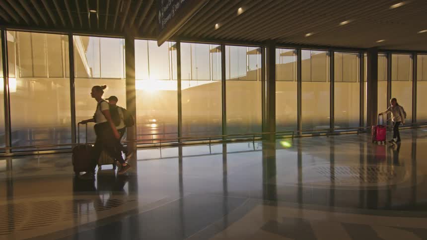 Travelers walking with luggage through a corridor lit by the rising sun at the airport