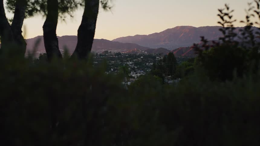 City surrounded by mountains is seen at sunset between trees