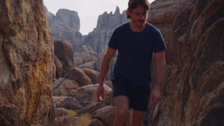 A man wearing a blue tshirt and black shorts walks up a rocky formation on a mountain at midday