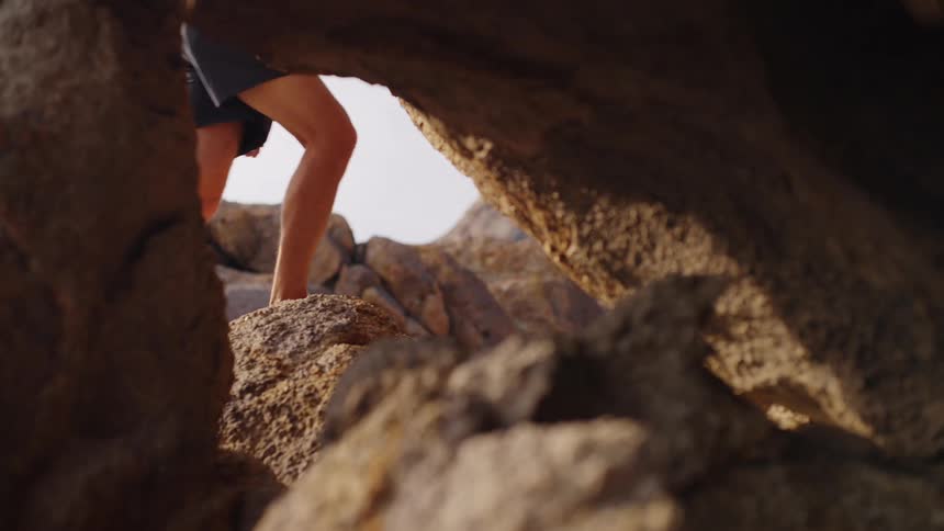 A low view between large brown rocks shows the legs of a person wearing dark shorts walking over the uneven rock surface with a bright sky in the background