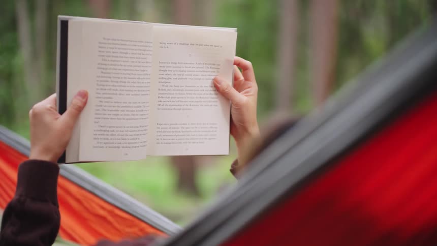 A person lying in a hammock near trees reading a book