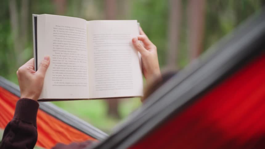 Person reading a book while resting in an orange hammock surrounded by nature