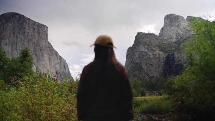 A woman in a baseball cap stands looking at the iconic granite cliffs of yosemite valley on a cloudy day