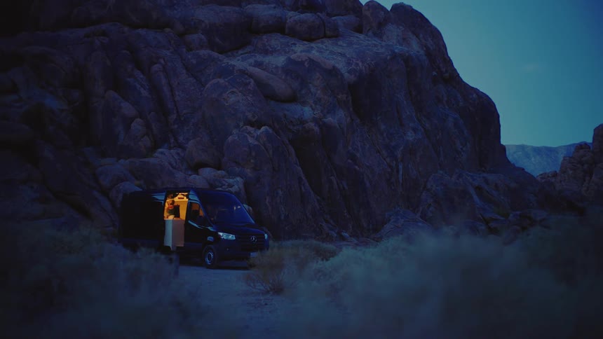 The video captures a darkcolored camper van parked off a dirt road in a rocky arid landscape at dusk or night the only illumination comes from the warm interior lights spilling out of the vans open side door contrasting with the dark rocks and deep