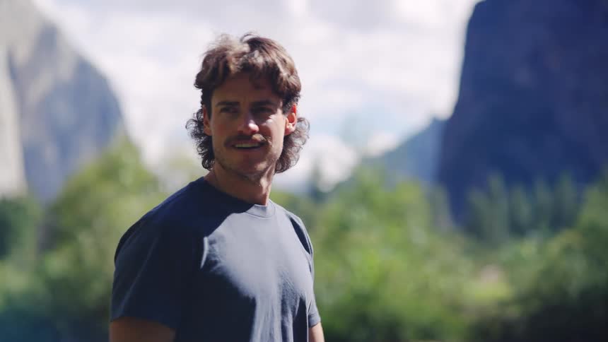 A man wearing a blue shirt stands in a lush green valley turns his head from profile to face forward with a slight smile and then turns away again with huge sharp mountain cliffs visible behind him