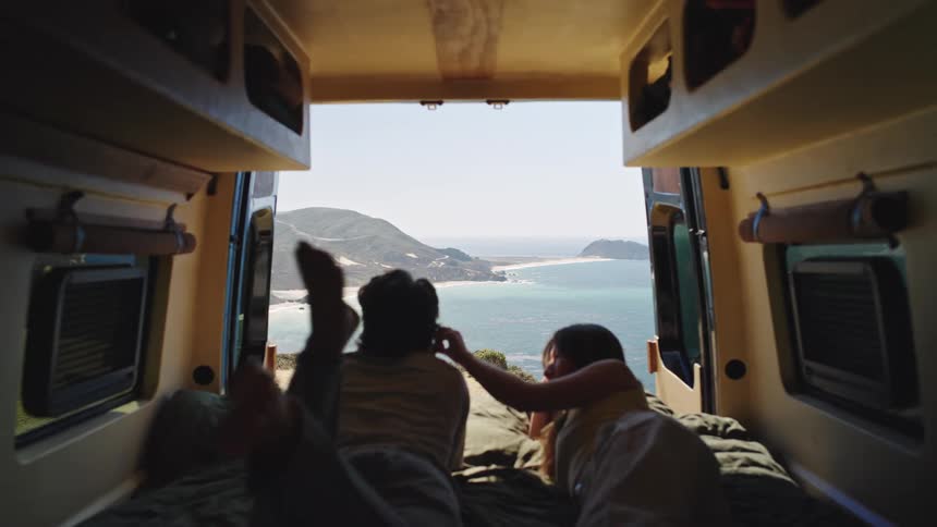 A young couple lies on a bed inside a camping van watching the ocean through the open back door as they engage in a discussion