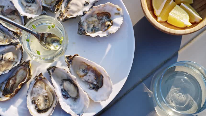 A hand reaches for oysters on a table beside lemon wedges in daylight