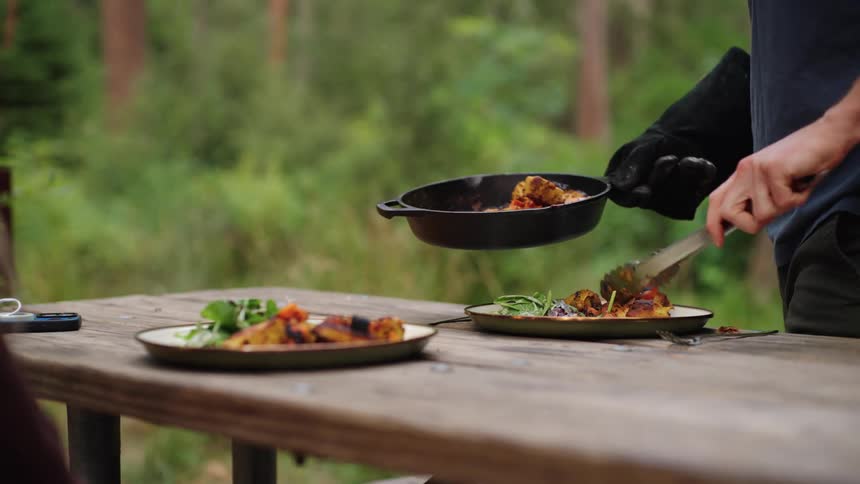 A person serves food onto plates at a wooden table in a forest