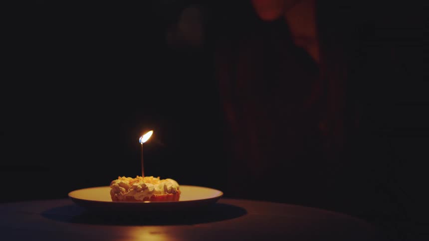 A hand places a white plate holding a small dessert with a lit candle onto a dark table a person leans into the frame and the candle flame is extinguished leaving the scene in deep shadow
