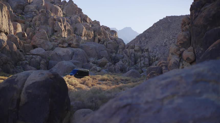 A wide view of a rugged desert landscape shows a small dark blue van driving through a boulder strewn valley framed by large rocks and dry brush with distant mountains visible