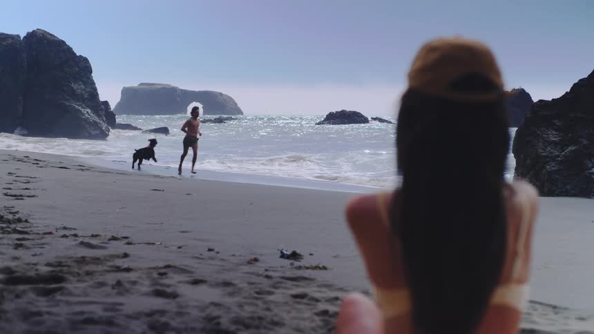 A man and a black dog run and play along the wet sand of a beach near the breaking waves viewed from behind a womans blurred shoulder and hair with large dramatic rock formations and a sea arch visible in the ocean background