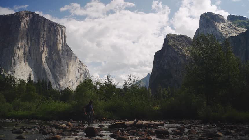 A man walks across rocky terrain passing a stream in the wild on a sunny day