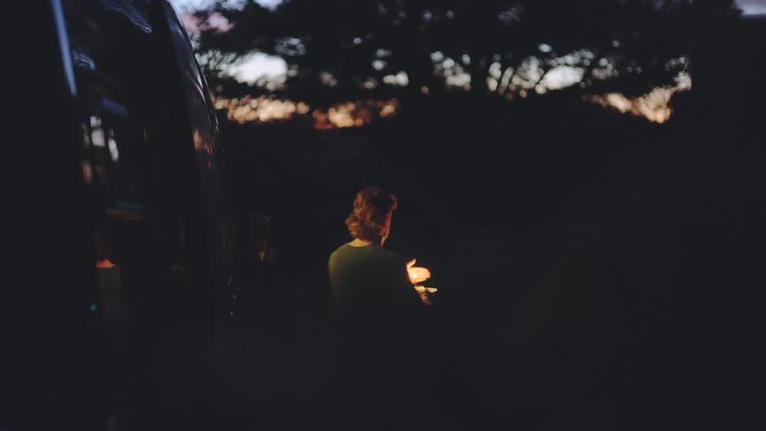 A young man steps out of a dark vehicle into a dark outdoor setting at dusk holding a bright flaming torch and walks away from the camera into the shadows