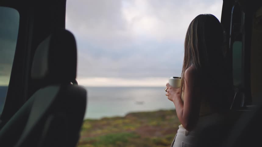 A woman sips coffee inside a van overlooking the ocean at dusk