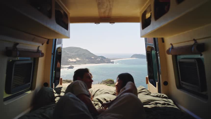Couple lying on bed inside van with open doors facing coastal landscape