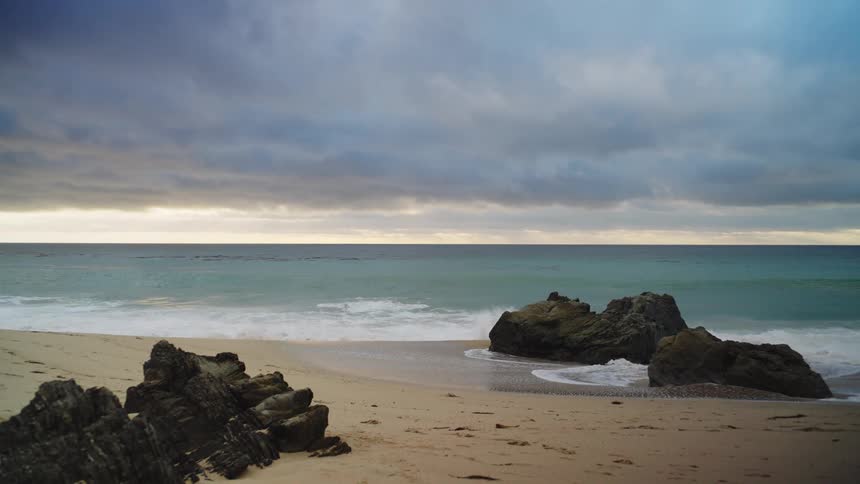 Waves crash against large rocks on a sandy beach under a cloudy sky